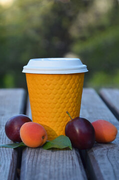 Disposible yellow paper cup with a white lid standing on  wooden table next to ripen plums and apricots. Closeup , center eyes level . Cup with beverage and fruits dessert on fresh air.