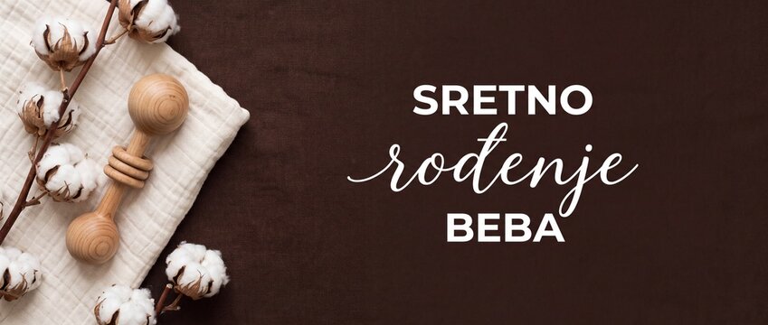 Baby shower newborn greeting card background featuring wooden rattle, organic cotton flowers, and text SRETNO RODENJE BEBA on dark brown fabric for newborn arrival announcement and congratulations.