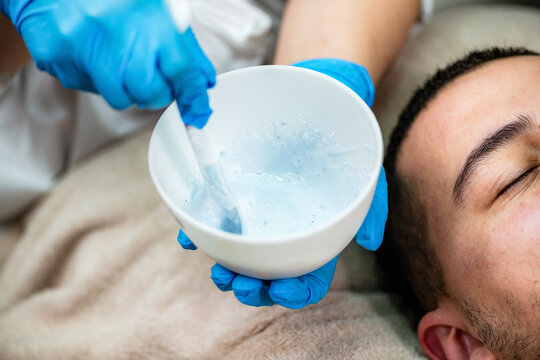Acne treatment for a young man receiving professional cosmetic facial care in beauty salon, with esthetician preparing a skincare mask for cleansing and blemish care.
