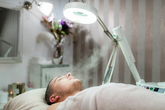 Young man relaxing during professional facial steaming treatment in beauty salon, focused on opening pores, skin cleansing, acne care and cosmetic skincare.