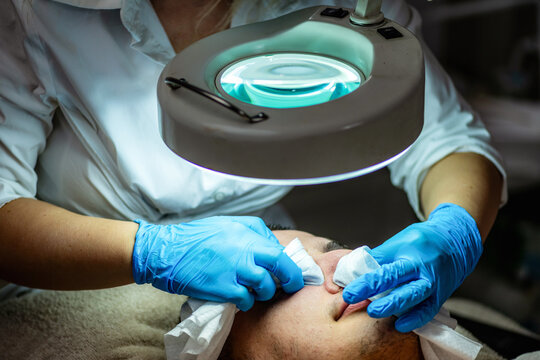 Young man receiving professional facial cleansing and acne care in beauty salon, with esthetician examining and treating the skin under a magnifying lamp.