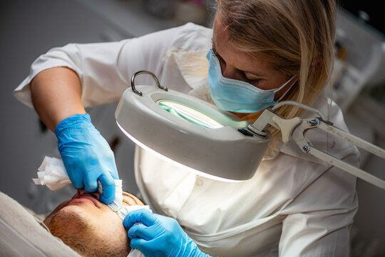 Professional acne treatment for a young man in beauty salon, with esthetician examining and cleansing facial skin under a cosmetic magnifying lamp.