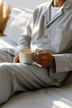 Close up on a man relaxing on a cozy sofa in soft grey striped pajamas and holding a mug of warm coffee or tea in the sunny morning.
