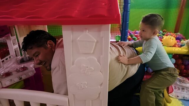 A funny scene featuring a toddler trying to pull his father out of a small plastic playhouse at an indoor children's playground with a ball pit in the background.