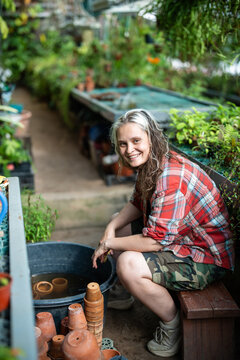 Cheerful smiling woman greenhouse employee sitting at orangery beside basin and ceramic pots, happy female plant nursery worker taking care of old terracotta pots, daily job in botanical garden.