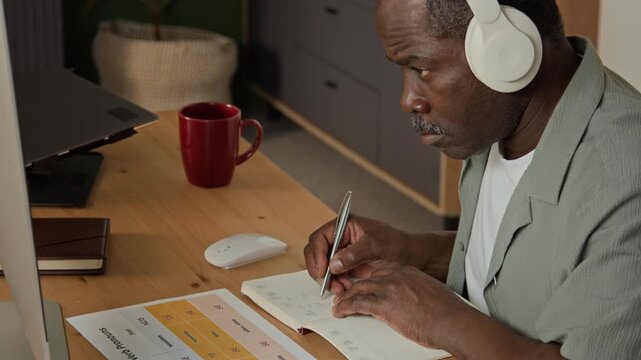 Mature African American man wearing headphones taking notes in notebook during online webinar at desk. Clip suitable for remote education and e-learning campaigns