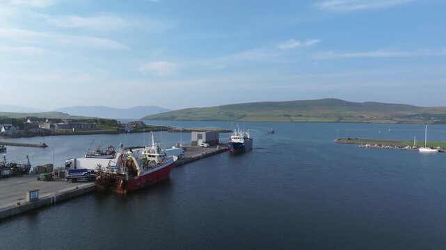 Fishing vessels line the concrete pier at Dingle Harbour as a larger ship steams out through the bay channel toward open water.