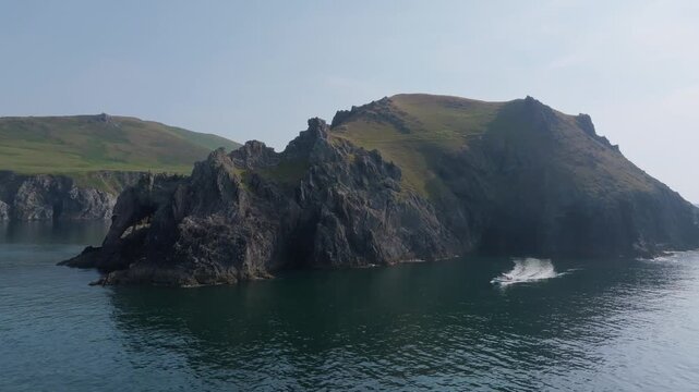 A small motorboat leaves a curved white wake as it passes jagged rock faces and grassy hills along the calm Dingle Peninsula coast.
