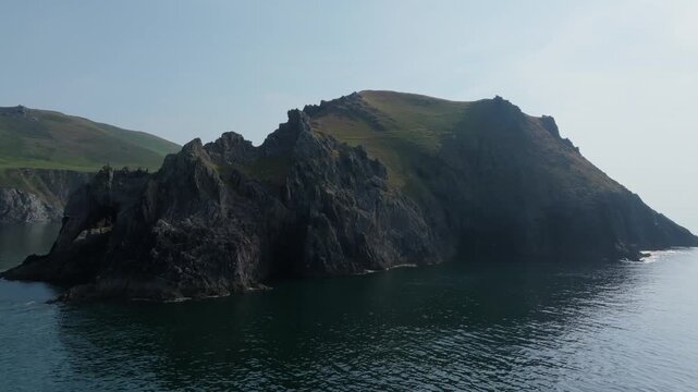Jagged dark cliffs rise from calm sea beside grass-covered slopes along the Dingle Peninsula coast, seen from a moving aerial viewpoint.