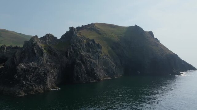 Steep rock faces drop into calm water beneath a broad grassy headland on the Dingle Peninsula, seen in a slowly moving aerial flyby.