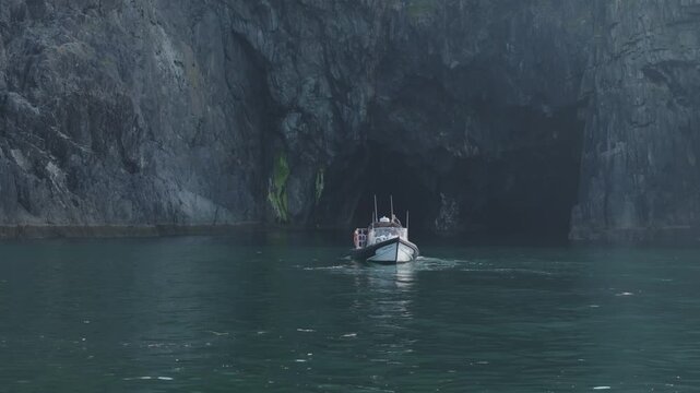 Small passenger boat moves toward a dark sea cave in steep rock walls, useful for adventure travel or coastal tourism themes.