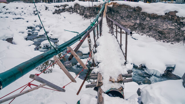 Makeshift wooden footbridge with metal railing crossing snowy mountain stream in rural village