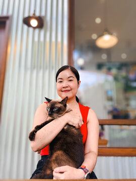 Portrait of a smiling Asian woman in a red top holding a brown Siamese cat outdoors with a blurred cafe background