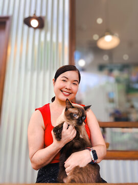 Portrait of a smiling Asian woman in a red top holding a brown Siamese cat outdoors with a blurred cafe background