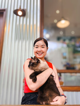 Portrait of a smiling Asian woman in a red top holding a brown Siamese cat outdoors with a blurred cafe background