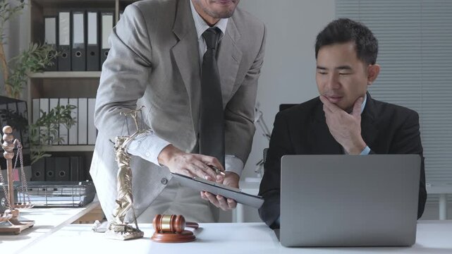 Asian male lawyers collaborating on a case using a laptop and documents in a modern law firm office with a gavel and justice statue
