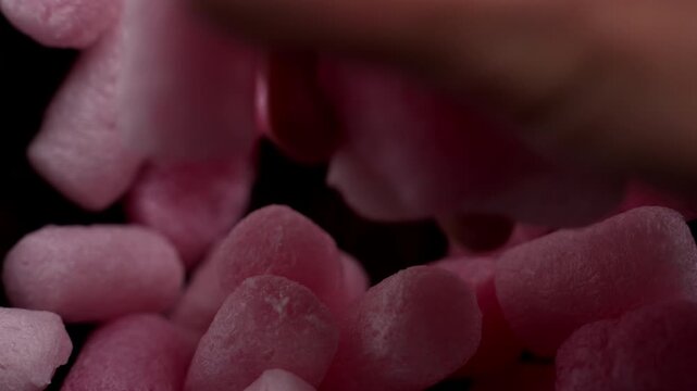 Hand picking up a pink packing peanut filler from a pile of lightweight protective packaging material. Fingers grasp porous irregularly shaped piece