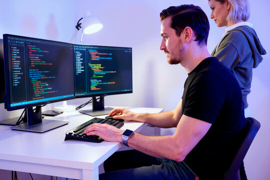 Caucasian young adult man writing code on dual monitors while Caucasian young adult woman stands nearby, representing software development teamwork, ideal for IT recruitment ads