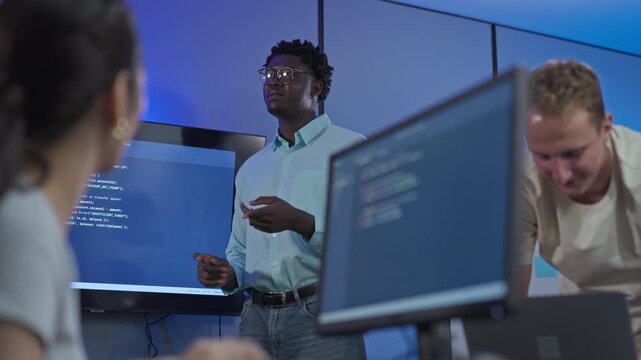 Young male programmer talks through transaction code on screen with colleagues while building banking and payment software at fintech company