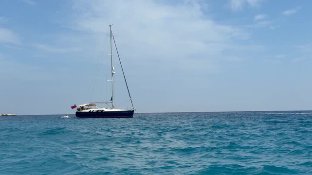 Sailboat cruises across open water beneath soft scattered clouds. Yacht moves slowly through mild waves near horizon, bringing relaxed feeling to sunny seascape, with sail, mast, and rigging visible.