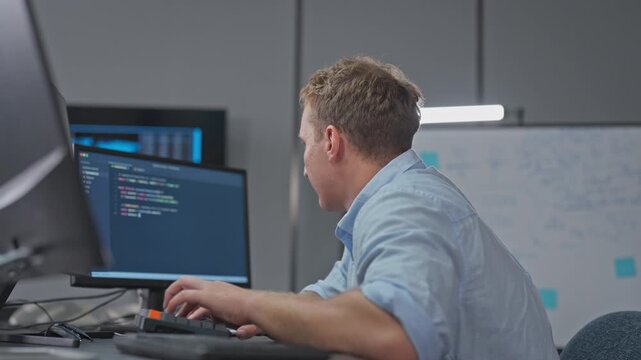 Young man types on his keyboard and reads code on his monitor, writing software at a quiet desk with a whiteboard behind him.