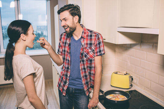 Lovely pictue of man standing in front of woman and holding wooden spoon with piece of omlet. She looks at it and keeps mouth opened. Girl is going to taste omlet. Guy is smiling. He is excited.
