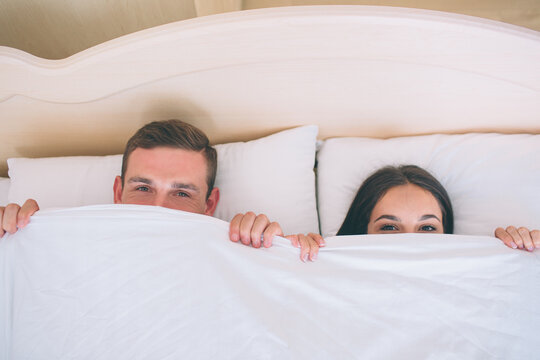 A picture of man and woman hiding faces under white blanket. They are lying together in one bed. Peple are looking up straight forward.