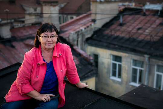Portrait of a woman wearing glasses and a pink jacket sitting on a rooftop with old city buildings in the background