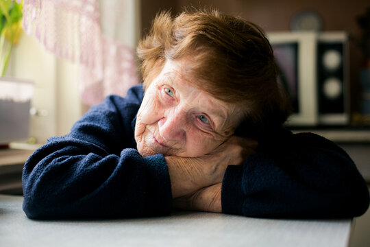 Close up portrait of an elderly woman resting her head on folded arms at a table by a window