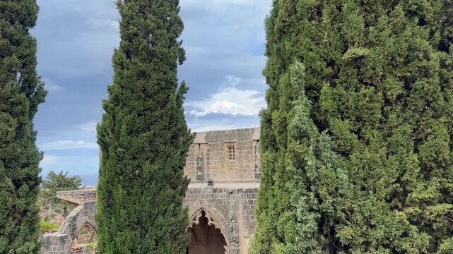 Kyrenia, Cyprus - October 3, 2026 Bellapais Abbey stands among tall cypress trees, with camera movement revealing Gothic arches and historic stonework. Old monastery overlooks landscaped grounds.