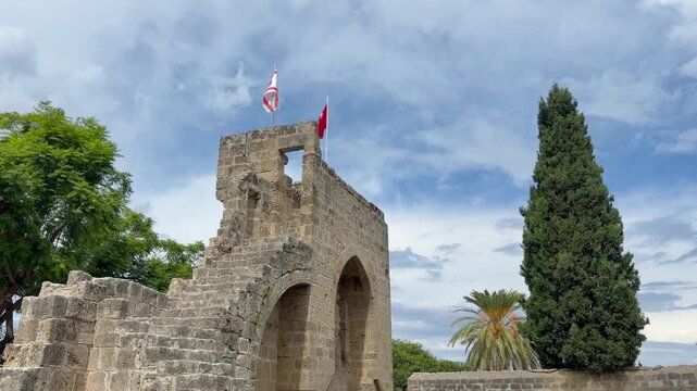 Kyrenia, Cyprus - October 3, 2026 Bellapais Abbey rises above leafy grounds, its Gothic stone arches and ruined tower framed by tall cypress and palm trees. Flags wave over historic masonry.