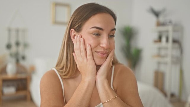 Woman cupping cheeks with both hands, smiling and looking aside inside a building bedroom set; calm selfcare wellbeing confidence.