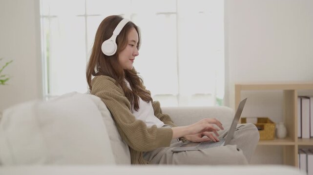 Young woman with headphone enjoying relaxation on sofa in home interior, brunette adult in casual clothing using laptop, beige decor, natural light, calm remote work and leisure lifestyle