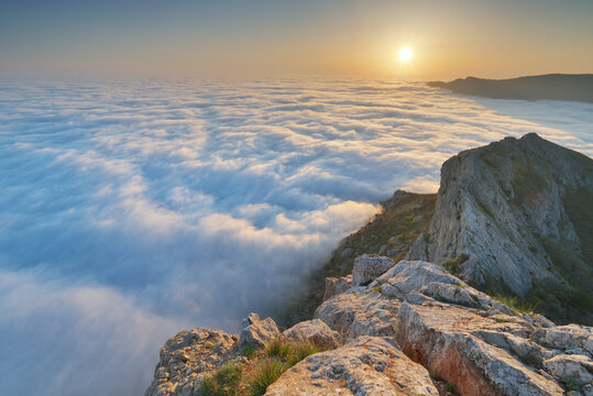 Rocky mountain peak rises above dense white cumulus clouds sea surface under bright sunrise sunburst sky. Beautiful misty wilderness landscape