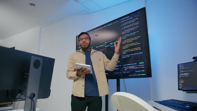Male DevOps Engineer Holding Digital Tablet and Explaining Software Deployment Architecture and Discussing Development Operations Strategy with Colleagues Reviewing Programming Code on Large Screen.