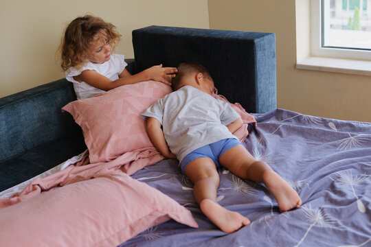 Caring little sister gently touching the head of her sleeping little brother lying on a large bed in a bright bedroom at home.