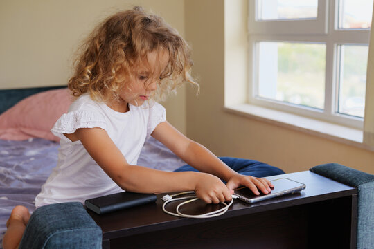 Cute little curly-haired girl connecting a USB cable to charge a smartphone with a portable power bank on a bedside table in a bright modern bedroom.