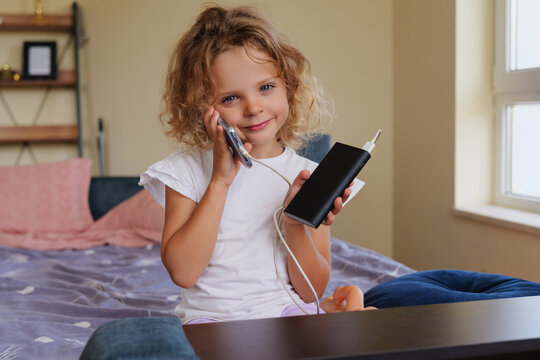Cute curly-haired little girl smiling while talking on a smartphone connected to a power bank, sitting on a bed in a cozy bedroom.
