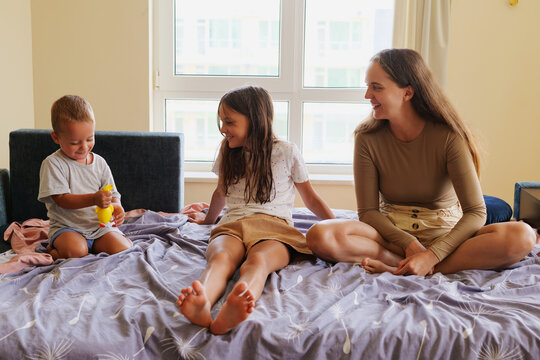 Happy mother and older daughter sitting on a bed, smiling and looking at a cute toddler boy playing with a toy in a bright bedroom.
