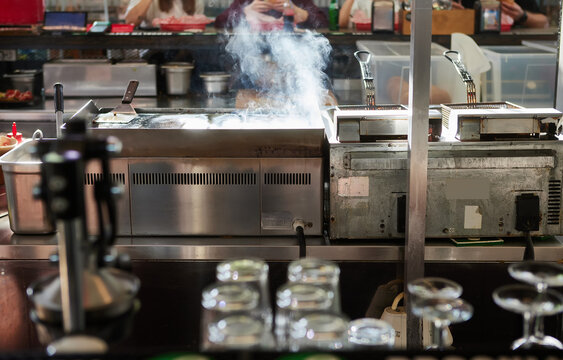 Commercial kitchen with grill, griddle and stainless steel cooking equipment. Steam rises from a hot cooking surface in a restaurant or food service environment, with glassware and staff 