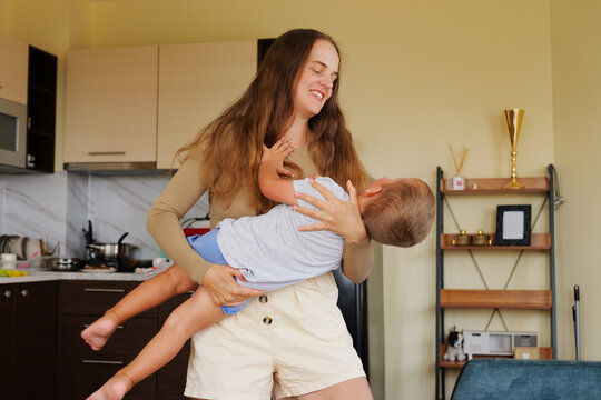 Happy mother holding and spinning her little toddler son in her arms in a modern bright kitchen at home.