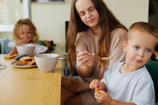 Mother trying to feed her stubborn little son with a spoon during family breakfast in a bright modern kitchen at home.