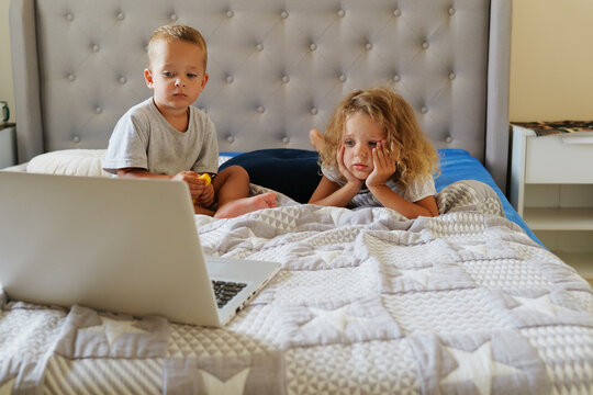 Young girl and her toddler brother focused on watching content on a laptop screen while lying and sitting on a bed in a cozy bedroom.