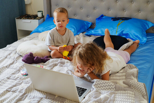 Young girl lying on a bed and watching a laptop screen while her little brother sits beside her holding a toy in a cozy bedroom.