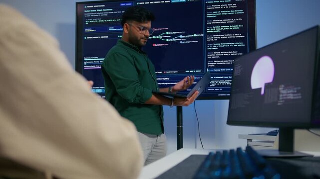 Male DevOps Engineer Holding Digital Tablet and Explaining Software Deployment Architecture and Discussing Development Operations Strategy with Colleagues Reviewing Programming Code on Large Screen.