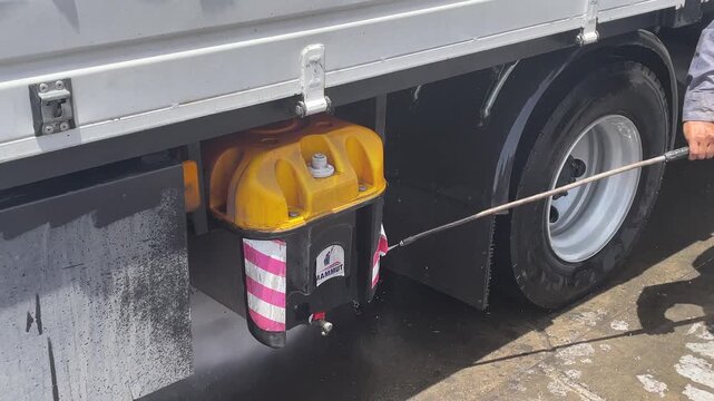 Water sprays beside a yellow container mounted under a truck cargo bed, with its holder, the rear tire and side frame visible in the washing area, highlighting truck care during maintenance.