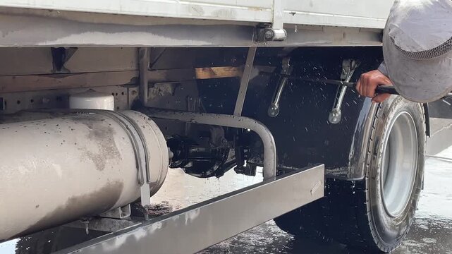 The lower side of a truck reveals a cylindrical tank, frame rail and underbody equipment, with water visible during a chassis washing session, illustrating truck cleaning for road transport.