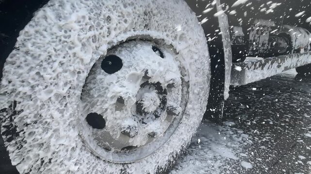 A spray of washing foam crosses a truck tire and metal rim, covering the wheel face with soap beneath the fender during exterior cleaning, highlighting transport maintenance at ground level.