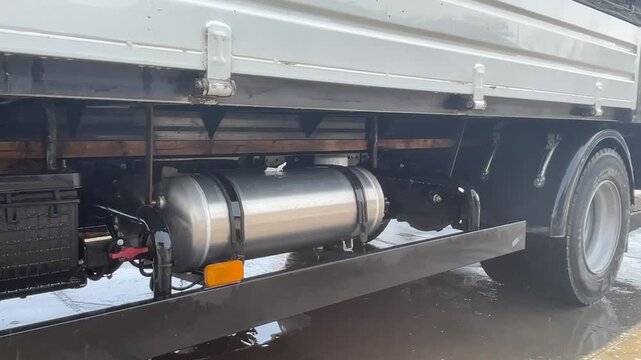 A metal tank is mounted along a truck chassis below the cargo bed, with nearby hoses, a storage box and a side rail visible above the wash floor, illustrating truck cleaning at ground level.