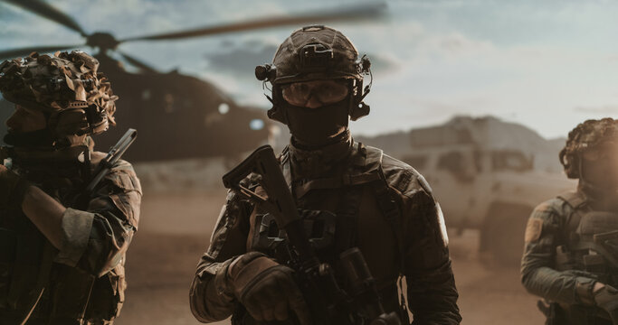 Close-up Portrait of a Masked Special Forces Operator in Helmet, Goggles and Headset Gripping His Carbine, Flanked by Teammates Beside a Helicopter and Armored Truck in Desert Dust.
